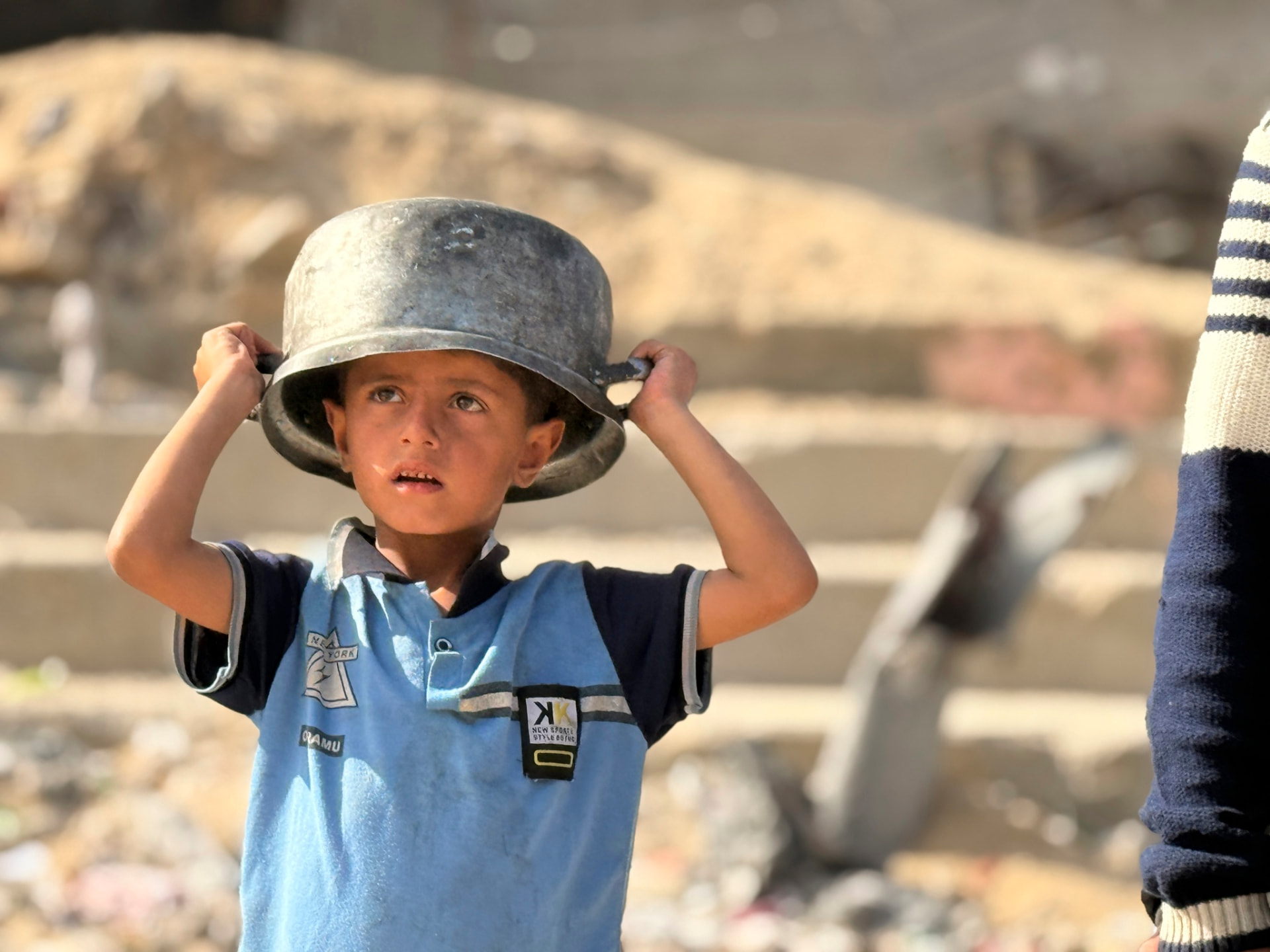 A boy in Gaza holds a dented cooking pot on his head, standing among rubble, waiting for aid